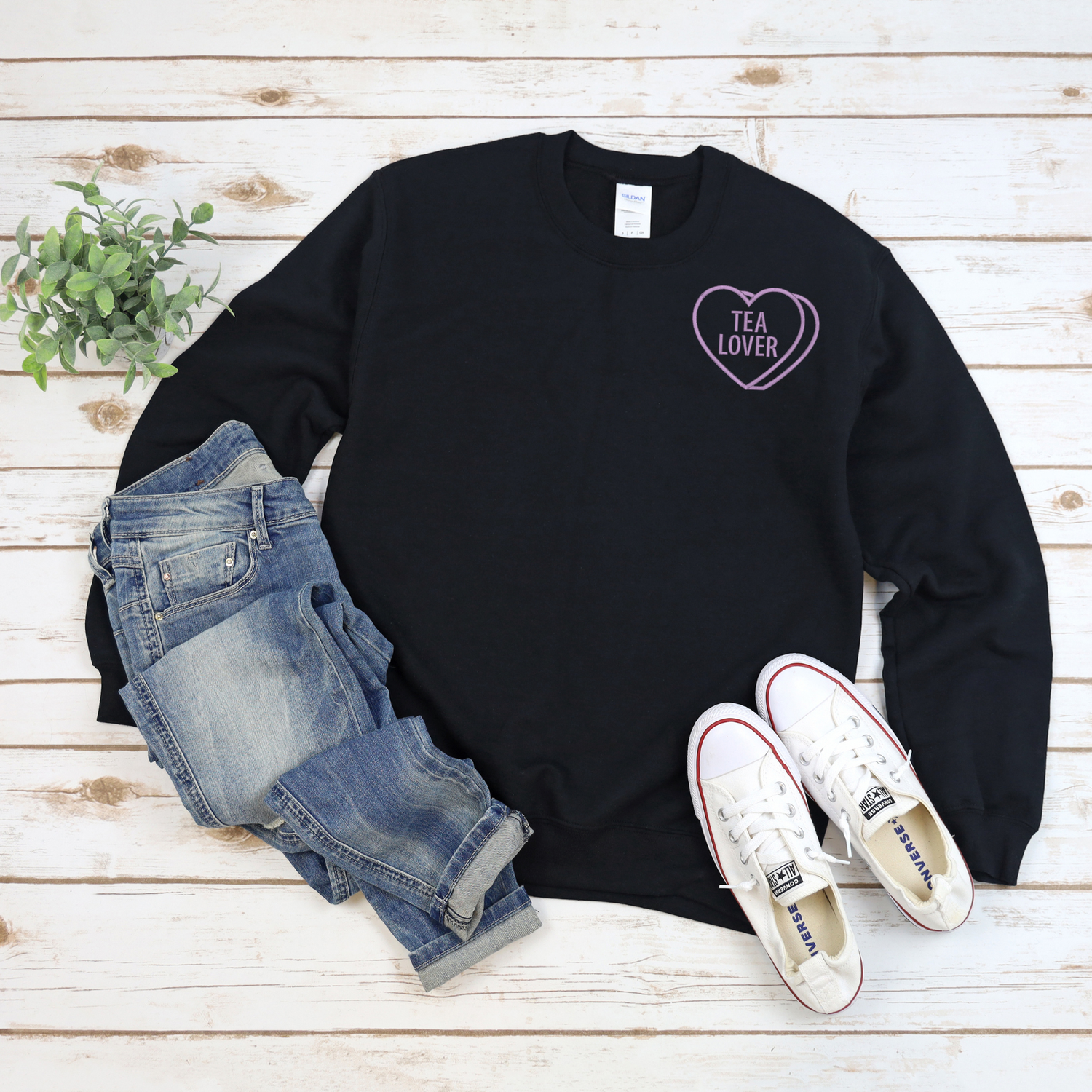 Black sweatshirt with 'TEA LOVER' embroidered in purple thread heart design, blue jeans, and white sneakers on a wooden surface.