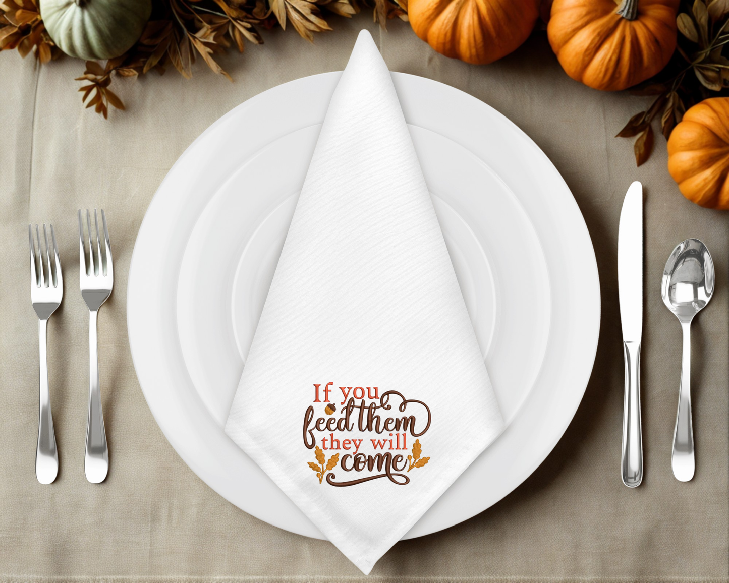 Thanksgiving table setting with a folded napkin featuring text "If you feed them they will come" and pumpkins in the background.